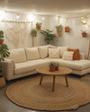 A cozy boho basement living area with a light cream sectional sofa, a round jute rug, and a natural wood coffee table. Warm lighting from a rattan floor lamp and string lights illuminates various potted plants, macrame wall hangings, and terracotta accents. — styling and décor detail
