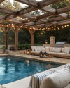A cozy farmhouse pool area featuring a large swimming pool with natural stone coping, surrounded by reclaimed wood pergola. Plush outdoor sofas with cream cushions and striped pillows are arranged on a flagstone patio, illuminated by Edison bulb string lights overhead and decorative lanterns.