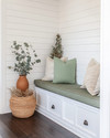 A bright farmhouse mudroom with white shiplap walls, a built-in bench with a sage green cushion, and dark wood flooring. Seasonal touches include a vase of eucalyptus, woven baskets, and a terracotta pot with dried botanicals, all bathed in warm, natural light. — styling and décor detail