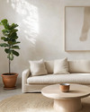 A serene Japandi great room featuring a low-slung oatmeal linen sofa, a light oak round coffee table, and a natural jute rug. Walls are painted in a soft off-white, adorned with a minimalist abstract nature print. A tall Fiddle Leaf Fig in a terracotta pot stands beside a ceramic table lamp with a linen shade, creating a calm and inviting atmosphere. — styling and décor detail