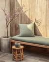 A Japandi porch blending vintage and modern, featuring a low-slung teak bench with a sage green linen cushion, a small hand-carved wooden stool as a side table, and a vintage Japanese stoneware vase with a single cherry blossom branch, all set against a backdrop of weathered wood and concrete planters. — styling and décor detail