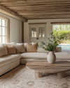 A cozy farmhouse poolhouse interior featuring a large cream sectional sofa with woven throw pillows, a distressed wood coffee table, and an exposed reclaimed wood ceiling. Soft light filters in, highlighting a natural fiber rug and a simple ceramic vase with greenery. — close-up of textures and materials