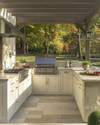 A refined traditional outdoor kitchen featuring off-white Shaker-style cabinetry, honed Pietra Cardosa countertops, and integrated stainless steel appliances under a simple pergola, with natural stone pavers. — alternate angle
