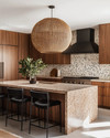 A Mid-Century Modern kitchen featuring flat-panel walnut cabinetry, a honed quartzite island, and a geometric terrazzo backsplash. A large woven rattan pendant light hangs above the island, which is flanked by black leather bar stools, all in soft natural daylight. — alternate angle