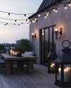 Industrial deck at dusk featuring warm white Edison bulb string lights draped overhead, dark bronze gooseneck wall sconces, a raw wood dining table, and large black metal lanterns with LED candles. — styling and décor detail