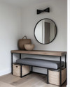 A modern mudroom features a natural oak console table with a minimalist black metal frame, paired with a charcoal grey upholstered bench. Overhead, a matte black sculptural flush-mount light illuminates the space, complemented by a large, frameless circular mirror above the console and woven jute baskets for storage. — close-up of textures and materials