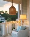 A well-lit coastal porch at dusk, featuring a woven rattan pendant light, string lights draped along the railing, and a floor lamp with a linen shade next to a white wicker chair with soft blue throw pillows. — close-up of textures and materials