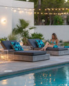 Modern poolside area with low-profile charcoal grey modular loungers, clear acrylic side tables, and large faux concrete planters. The scene is illuminated by sleek rechargeable LED lanterns and subtle solar-powered string lights, with deep teal and slate blue geometric outdoor cushions adding pops of color.