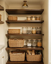 A cozy traditional pantry featuring warm cream walls, rich dark wood shelving, and a mix of woven rattan baskets and clear glass jars holding pantry staples. An aged brass semi-flush mount light fixture illuminates the space. — alternate angle