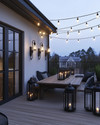 Industrial deck at dusk featuring warm white Edison bulb string lights draped overhead, dark bronze gooseneck wall sconces, a raw wood dining table, and large black metal lanterns with LED candles. — alternate angle