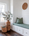 A bright farmhouse mudroom with white shiplap walls, a built-in bench with a sage green cushion, and dark wood flooring. Seasonal touches include a vase of eucalyptus, woven baskets, and a terracotta pot with dried botanicals, all bathed in warm, natural light. — alternate angle