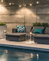 Modern poolside area with low-profile charcoal grey modular loungers, clear acrylic side tables, and large faux concrete planters. The scene is illuminated by sleek rechargeable LED lanterns and subtle solar-powered string lights, with deep teal and slate blue geometric outdoor cushions adding pops of color. — close-up of textures and materials