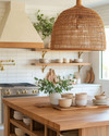 A bohemian kitchen featuring a large woven rattan pendant light above a wooden island with fresh greenery, open shelving with ceramic dishes, and warm brass fixtures creating a cozy, inviting ambiance. — styling and décor detail