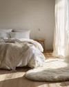 A cozy Scandinavian bedroom featuring a bed dressed in white linen, a chunky knit throw, and a sheepskin rug on light wooden floorboards. Soft, diffused light comes from a simple bedside lamp and a large window. — close-up of textures and materials