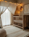 A rustic baby and kids room with a warm, soft glow from layered lighting, featuring a natural wood crib, a cream wool rug, linen curtains, and subtle string lights draped over a canopy. — alternate angle