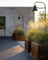 An industrial driveway featuring dark gray sealed concrete, bordered by robust steel edging, with matte black gooseneck lighting fixtures mounted on a raw concrete wall and large Corten steel planters filled with hardy ornamental grasses. — styling and décor detail