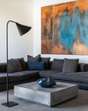 A modern family room featuring a large charcoal grey sectional sofa, a sculptural concrete coffee table, and a large abstract painting with blues and sienna tones. A tall, slender black metal floor lamp stands next to the sofa. — styling and décor detail
