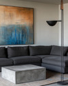 A modern family room featuring a large charcoal grey sectional sofa, a sculptural concrete coffee table, and a large abstract painting with blues and sienna tones. A tall, slender black metal floor lamp stands next to the sofa. — close-up of textures and materials