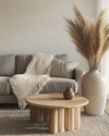 A cozy modern landscape living room featuring a warm gray sofa, a chunky cream knit throw, a light oak coffee table, and a large ceramic vase with dried pampas grass, all illuminated by soft natural light. — styling and décor detail