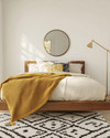 A renter-friendly Mid-Century Modern bedroom featuring a walnut platform bed with a cream duvet, a mustard yellow knit throw, and a graphic black and white area rug. Brass multi-arm floor lamp and a simple round mirror accent the space. — alternate angle