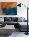 A modern family room featuring a large charcoal grey sectional sofa, a sculptural concrete coffee table, and a large abstract painting with blues and sienna tones. A tall, slender black metal floor lamp stands next to the sofa. — alternate angle