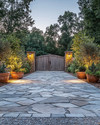 Rustic driveway with warm grey gravel and large, irregular flagstones leading to a wooden gate. The scene is enhanced by low-level pathway lights, subtle uplighting on surrounding trees, and terracotta planters filled with native greenery, all under a soft, twilight glow.