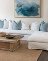 A serene coastal family room featuring a large linen slipcovered sectional sofa, a reclaimed wood coffee table, and a woven jute rug. Soft blue and white throw pillows add subtle color, complemented by a large abstract ocean-inspired artwork above the sofa and natural rattan accents.