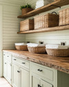 A cozy farmhouse laundry room featuring reclaimed wood countertops, open wooden shelving, and large wicker laundry baskets. Walls are painted in a soft sage green, complemented by creamy white shiplap, and matte black hardware on cabinets. — styling and décor detail