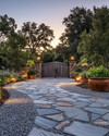 Rustic driveway with warm grey gravel and large, irregular flagstones leading to a wooden gate. The scene is enhanced by low-level pathway lights, subtle uplighting on surrounding trees, and terracotta planters filled with native greenery, all under a soft, twilight glow. — styling and décor detail