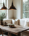 A warm and inviting farmhouse breakfast nook featuring a rustic reclaimed wood table, a cream banquette with linen cushions, and two aged bronze pendant lights hanging over the table, with soft morning light streaming through a window. — styling and décor detail