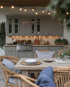 Cozy coastal outdoor kitchen featuring weathered gray cabinetry, honed travertine countertops, a teak dining table with comfortable cushioned chairs, and warm string lights overhead, accented with woven rattan and soft indigo textiles. — close-up of textures and materials