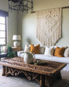 A Farmhouse Craft living room featuring a large, hand-carved reclaimed oak coffee table, a woven macrame wall hanging in ivory, and a wrought-iron chandelier, with ceramic table lamps and deep ochre throw pillows.