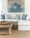 A serene coastal family room featuring a large linen slipcovered sectional sofa, a reclaimed wood coffee table, and a woven jute rug. Soft blue and white throw pillows add subtle color, complemented by a large abstract ocean-inspired artwork above the sofa and natural rattan accents. — close-up of textures and materials