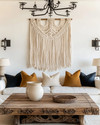 A Farmhouse Craft living room featuring a large, hand-carved reclaimed oak coffee table, a woven macrame wall hanging in ivory, and a wrought-iron chandelier, with ceramic table lamps and deep ochre throw pillows. — close-up of textures and materials