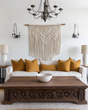 A Farmhouse Craft living room featuring a large, hand-carved reclaimed oak coffee table, a woven macrame wall hanging in ivory, and a wrought-iron chandelier, with ceramic table lamps and deep ochre throw pillows. — alternate angle