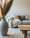 A cozy modern landscape living room featuring a warm gray sofa, a chunky cream knit throw, a light oak coffee table, and a large ceramic vase with dried pampas grass, all illuminated by soft natural light. — alternate angle
