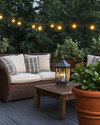 A cozy farmhouse deck features deep-seated wicker furniture with cream cushions and plaid throw pillows, illuminated by warm string lights and a metal lantern. Terracotta pots with lush greenery and a rustic wooden coffee table complete the inviting outdoor scene.