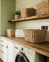 A cozy farmhouse laundry room featuring reclaimed wood countertops, open wooden shelving, and large wicker laundry baskets. Walls are painted in a soft sage green, complemented by creamy white shiplap, and matte black hardware on cabinets. — close-up of textures and materials
