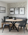 A modern breakfast nook features a light wood round table with a minimalist black metal base, paired with a charcoal grey upholstered bench and two black metal chairs. A subtle geometric peel-and-stick wallpaper adorns the wall, alongside a gallery of abstract art prints.