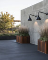 An industrial driveway featuring dark gray sealed concrete, bordered by robust steel edging, with matte black gooseneck lighting fixtures mounted on a raw concrete wall and large Corten steel planters filled with hardy ornamental grasses. — alternate angle