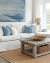 A serene coastal family room featuring a large linen slipcovered sectional sofa, a reclaimed wood coffee table, and a woven jute rug. Soft blue and white throw pillows add subtle color, complemented by a large abstract ocean-inspired artwork above the sofa and natural rattan accents. — styling and décor detail
