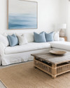 A serene coastal family room featuring a large linen slipcovered sectional sofa, a reclaimed wood coffee table, and a woven jute rug. Soft blue and white throw pillows add subtle color, complemented by a large abstract ocean-inspired artwork above the sofa and natural rattan accents. — alternate angle