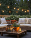 A cozy farmhouse deck features deep-seated wicker furniture with cream cushions and plaid throw pillows, illuminated by warm string lights and a metal lantern. Terracotta pots with lush greenery and a rustic wooden coffee table complete the inviting outdoor scene. — styling and décor detail