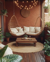 A cozy boho porch featuring a rattan hanging chair, a jute rug, and a low wooden coffee table surrounded by plush cream cushions. Fairy lights are strung overhead, and various potted plants add greenery against a backdrop of warm terracotta walls. — styling and décor detail