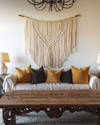 A Farmhouse Craft living room featuring a large, hand-carved reclaimed oak coffee table, a woven macrame wall hanging in ivory, and a wrought-iron chandelier, with ceramic table lamps and deep ochre throw pillows. — styling and décor detail
