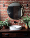 Industrial bathroom with a warm red exposed brick wall, a dark wood reclaimed vanity with a white ceramic basin, and matte black fixtures. A large round mirror with an aged brass frame hangs above the vanity, complemented by a lush green fern in a terracotta pot. — styling and décor detail