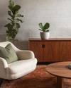 A Mid-Century Modern living room featuring a rich walnut credenza, a teak coffee table, and a cream bouclé lounge chair. The space is anchored by a wool rug in deep terracotta, accented with sage green linen pillows, and houses a tall fiddle-leaf fig in an unglazed ceramic planter. — close-up of textures and materials