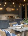 Cozy coastal outdoor kitchen featuring weathered gray cabinetry, honed travertine countertops, a teak dining table with comfortable cushioned chairs, and warm string lights overhead, accented with woven rattan and soft indigo textiles. — styling and décor detail