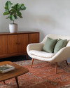 A Mid-Century Modern living room featuring a rich walnut credenza, a teak coffee table, and a cream bouclé lounge chair. The space is anchored by a wool rug in deep terracotta, accented with sage green linen pillows, and houses a tall fiddle-leaf fig in an unglazed ceramic planter. — styling and décor detail