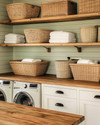 A cozy farmhouse laundry room featuring reclaimed wood countertops, open wooden shelving, and large wicker laundry baskets. Walls are painted in a soft sage green, complemented by creamy white shiplap, and matte black hardware on cabinets. — alternate angle